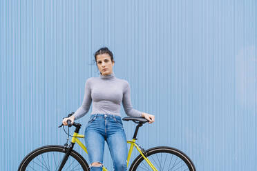 Selbstbewusste Frau im mittleren Erwachsenenalter sitzt auf einem Fixie-Fahrrad vor einer blauen Wand - DAMF00645