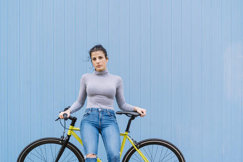 Confident mid adult woman sitting on fixie bike against blue wall - DAMF00645