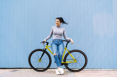 Thoughtful woman smiling while sitting on fixie bike against blue wall - DAMF00646