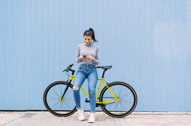 Lächelnde Frau, die ein Mobiltelefon benutzt, während sie auf einem Fixie-Fahrrad vor einer blauen Wand sitzt - DAMF00647