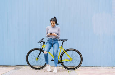 Smiling woman using mobile phone while sitting on fixie bike against blue wall - DAMF00647