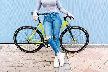 Mid adult woman wearing torn jeans sitting on fixie bike against blue wall - DAMF00649