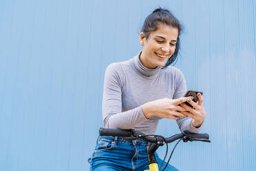 Fröhliche schöne Frau, die ein Mobiltelefon benutzt, während sie auf einem Fixie-Fahrrad vor einer blauen Wand sitzt - DAMF00650