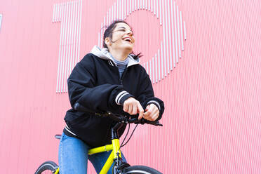 Fröhlich Mitte erwachsene Frau lachend, während auf fixie Fahrrad gegen rosa Wand sitzen - DAMF00651
