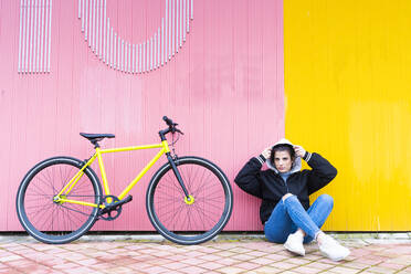 Schöne Frau mit Kapuze sitzt mit einem Fixie-Fahrrad auf der Straße an der Wand - DAMF00652