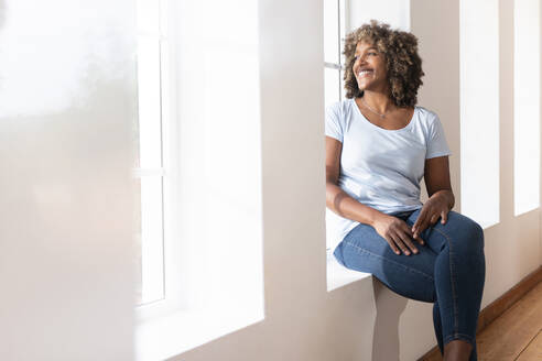 Mid adult woman looking away while sitting on window sill at home - SBOF02450