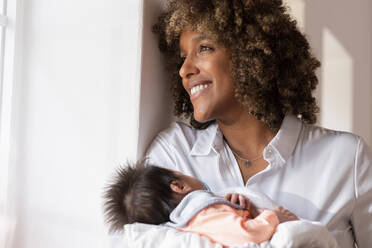 Happy mother with newborn baby looking away while standing by window at home - SBOF02457