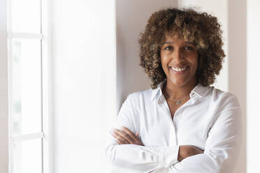 Curly hair woman standing with arms crossed by window at home - SBOF02462