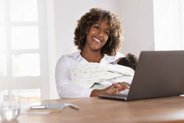 Smiling mother holding baby while working on laptop at home office - SBOF02468