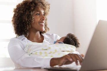 Smiling woman working on laptop while sitting with baby at home office - SBOF02471