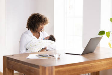 Mother looking at sleeping baby while sitting by table at home office - SBOF02474