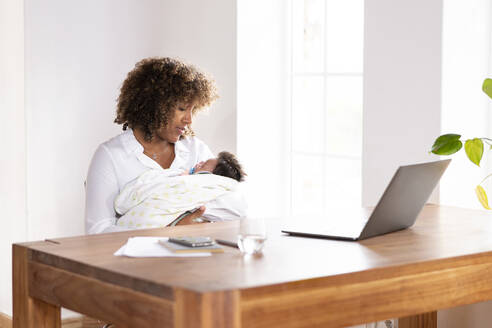 Mutter sieht schlafendes Baby an, während sie am Tisch im Büro zu Hause sitzt - SBOF02474