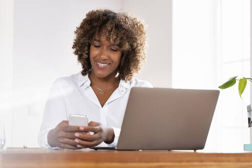 Geschäftsfrau mit Laptop und Mobiltelefon im Home Office sitzend - SBOF02482