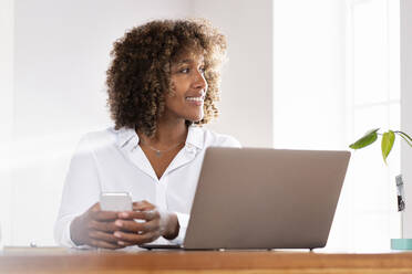 Smiling businesswoman with laptop and mobile phone looking away while sitting at home office - SBOF02484