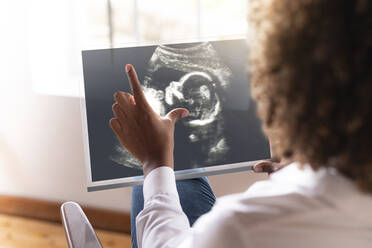 Frau betrachtet Ultraschallbild des Babys auf transparentem Bildschirm, während sie zu Hause sitzt - SBOF02486