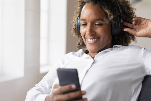 Lächelnde Frau, die Musik hört, während sie ein Mobiltelefon benutzt und zu Hause sitzt - SBOF02501