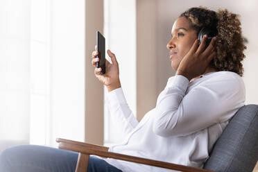Mittlere erwachsene Frau mit Mobiltelefon hört Musik über Kopfhörer, während sie zu Hause auf einem Sessel sitzt - SBOF02502
