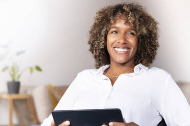 Lächelnde Frau mit digitalem Tablet, die wegschaut, während sie zu Hause sitzt - SBOF02516