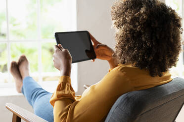 Frau mit lockigem Haar, die ein digitales Tablet benutzt, während sie sich auf einem Sessel zu Hause ausruht - SBOF02519