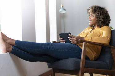 Lächelnde Frau mit digitalem Tablet, die wegschaut, während sie sich auf einem Sessel zu Hause entspannt - SBOF02527