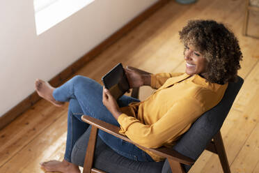 Lächelnde Frau mit digitalem Tablet, die wegschaut, während sie sich auf einem Sessel zu Hause ausruht - SBOF02528