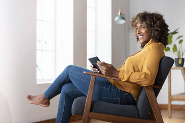 Mid adult woman with digital tablet looking away while sitting on armchair at home - SBOF02531