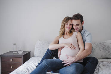 Lächelnder Mann berührt den Bauch seiner schwangeren Frau, während er zu Hause auf dem Bett sitzt - EBBF02233