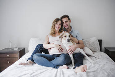 Lächelndes Paar mit Hund, das sich zu Hause auf dem Bett an der Wand entspannt - EBBF02236