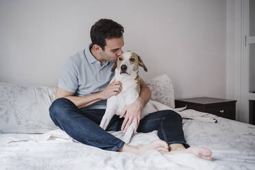 Mid adult man kissing dog while sitting on bed against wall at home - EBBF02237