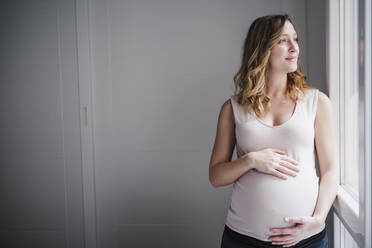 Thoughtful pregnant woman with hands on stomach looking through window at home - EBBF02239