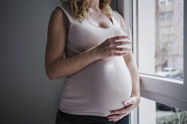 Pregnant woman holding drinking glass while standing by window at home - EBBF02240