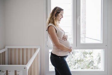 Schwangere Frau mit Händen auf dem Bauch nachdenklich, während sie zu Hause am Fenster steht - EBBF02242