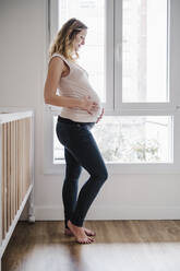 Schwangere Mutter mit Händen auf dem Bauch in Erwartung am Fenster stehend - EBBF02243