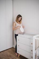 Smiling pregnant woman holding baby clothing while standing by cabinet at home - EBBF02246