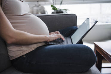 Pregnant woman using laptop while sitting on sofa at home - EBBF02250