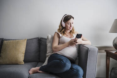 Schwangere Frau hört Musik über Kopfhörer, während sie auf dem Sofa zu Hause ihr Handy benutzt - EBBF02254