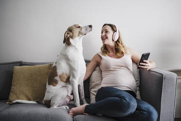 Pregnant mother listening music over headphones while sitting with dog on sofa at home - EBBF02255