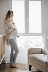 Schwangere Frau mit Händen auf dem Bauch steht am Fenster - EBBF02258