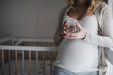 Close-up of pregnant woman holding drinking glass while leaning on crib at home - EBBF02259