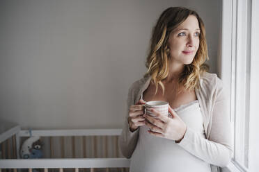 Nachdenkliche schwangere Frau, die eine Kaffeetasse hält, während sie zu Hause an der Krippe steht - EBBF02260
