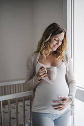 Smiling pregnant woman with hand on stomach holding coffee while standing at home - EBBF02261