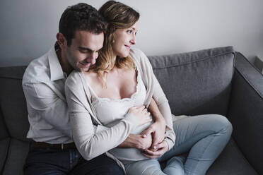 Mann mit geschlossenen Augen, der seine schwangere Frau umarmt, während er zu Hause auf dem Sofa sitzt - EBBF02262