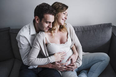 Ehemann, der seine schwangere Frau umarmt, während er zu Hause auf dem Sofa sitzt - EBBF02263