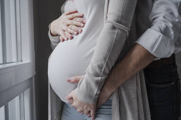 Pregnant woman with husband's hands on stomach standing by window at home - EBBF02264