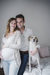 Lächelnder Mann mit schwangerer Frau und Hund im Schlafzimmer stehend - EBBF02266
