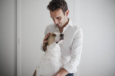 Mid adult man petting dog while standing against white wall at home - EBBF02269