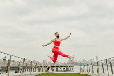 Junge Sportlerin springt in der Luft und trägt eine Gesichtsmaske am Ufer - CAVF91911