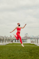 Young female athlete jumping mid air wearing face mask by waterfront. - CAVF91912