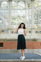 Smiling young woman wearing green skirt posing against greenhouse - JMPF00811