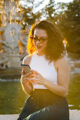 Frau mit Sonnenbrille, die ihr Smartphone benutzt, während sie an einem Brunnen im Park sitzt - JMPF00818